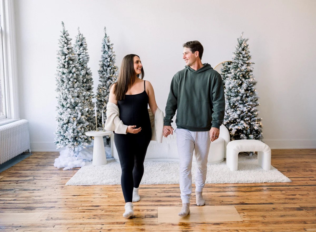 Expecting couple in coordinated neutral and green outfits walking through a snowy holiday studio set in Chicago